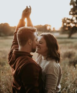A couple laughs together in a golden field, holding hands high.