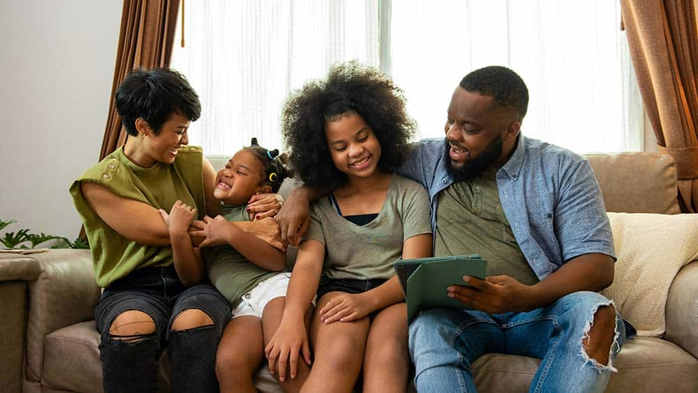 A family of four sits together on a couch, smiling as the father holds a tablet.