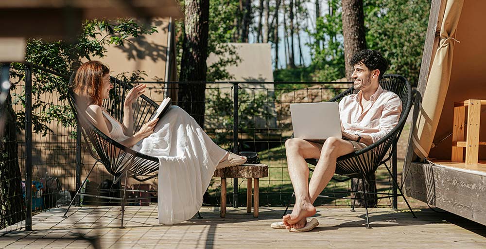 A man and woman relax in black woven chairs on a wooden deck outside a glamping tent.
