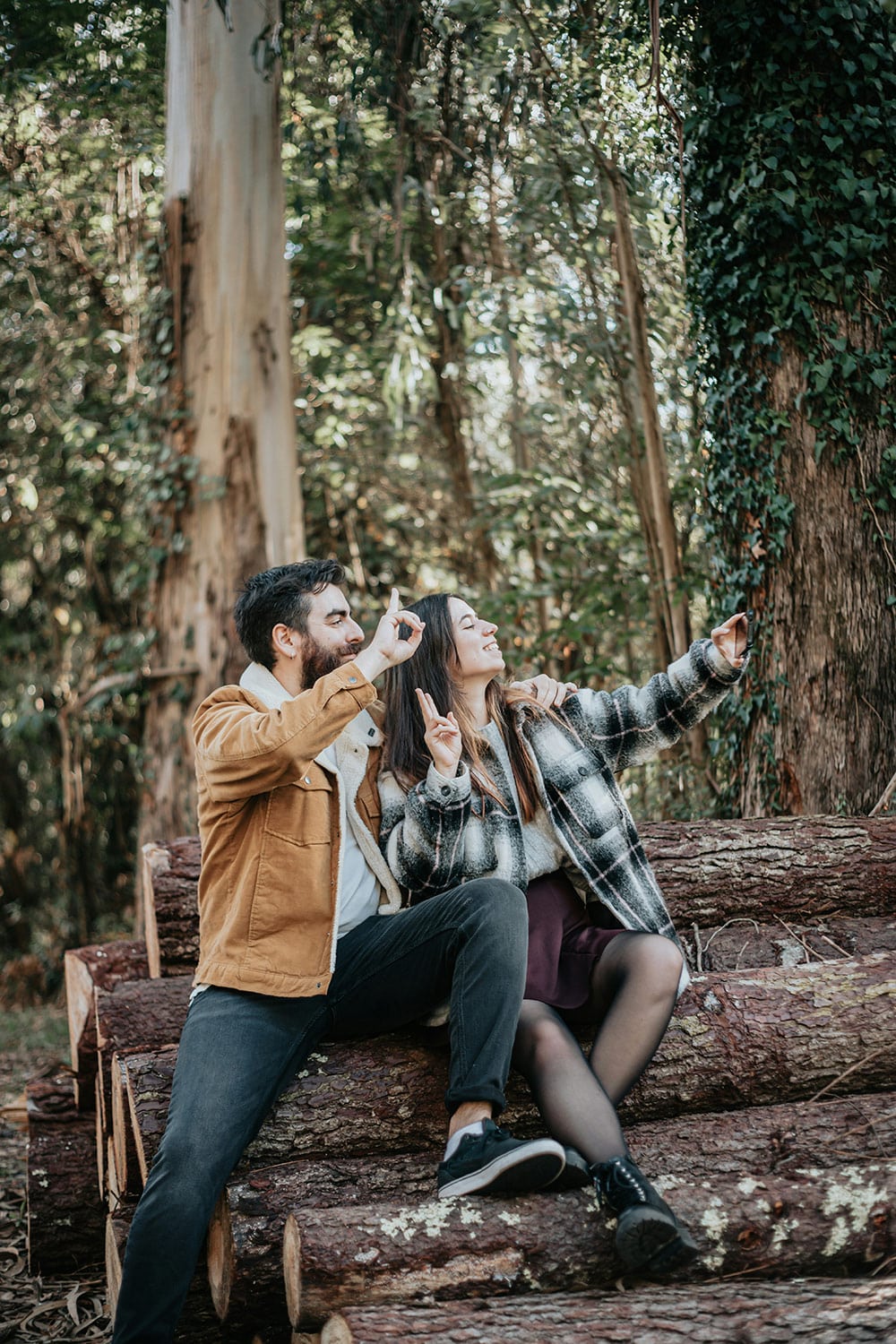 A man and woman sit on a stack of logs in a forest