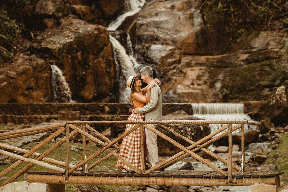A smiling couple embraces on a rustic wooden bridge with a scenic waterfall in the background.