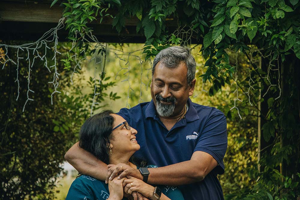 A smiling couple embraces outdoors, framed by lush green foliage and decorative string lights hanging from a wooden structure.