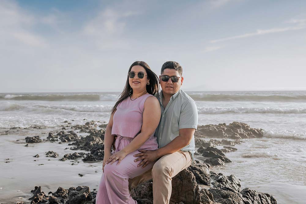 A smiling couple wearing sunglasses sits together on a rocky beach with ocean waves in the background.