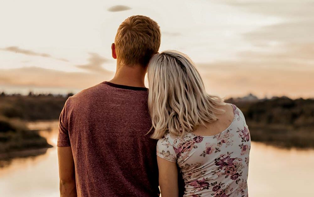 A young couple stands with their backs to the camera, looking out over a calm body of water at sunset.