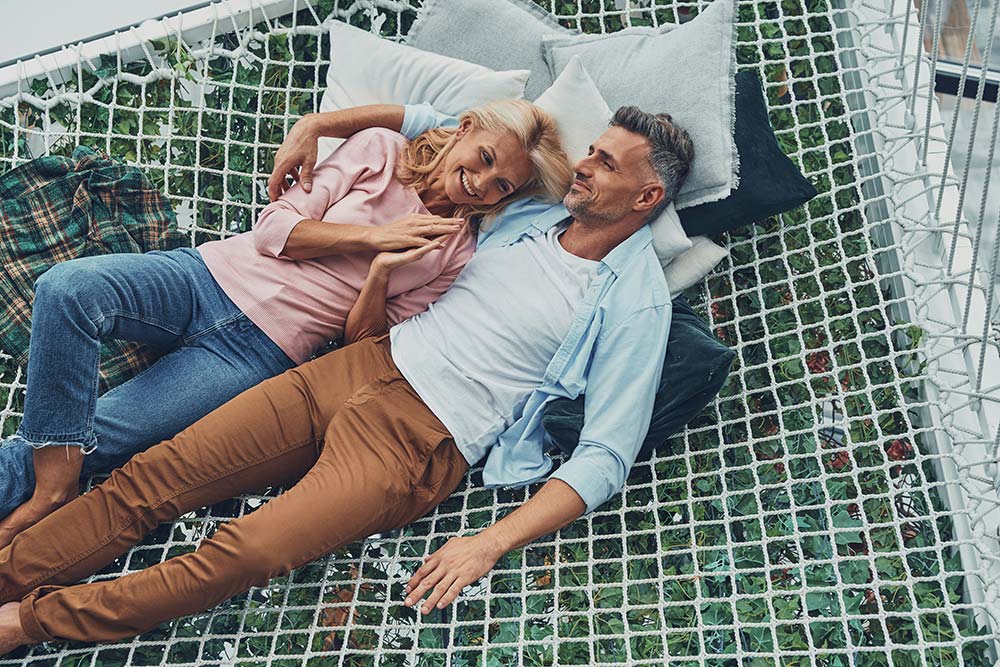 Top view of a mature couple smiling and talking while relaxing together in a large hammock at home.
