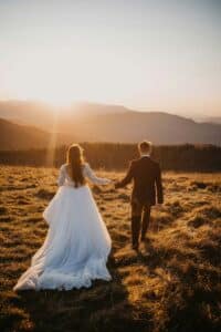 bride and groom holding hands and walking through a field at sunset.