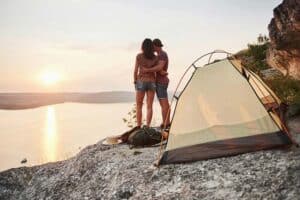 couple hugging next to a tent on a cliff overlooking a lake at sunset.