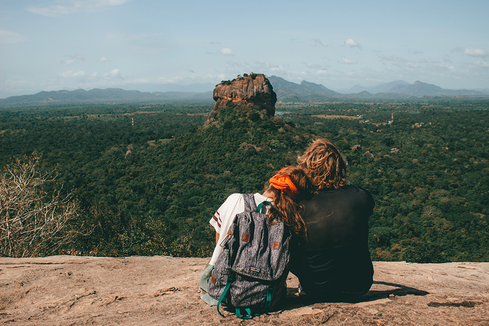 This image features a couple sitting side-by-side on a rocky ledge, overlooking a vast, lush green landscape.