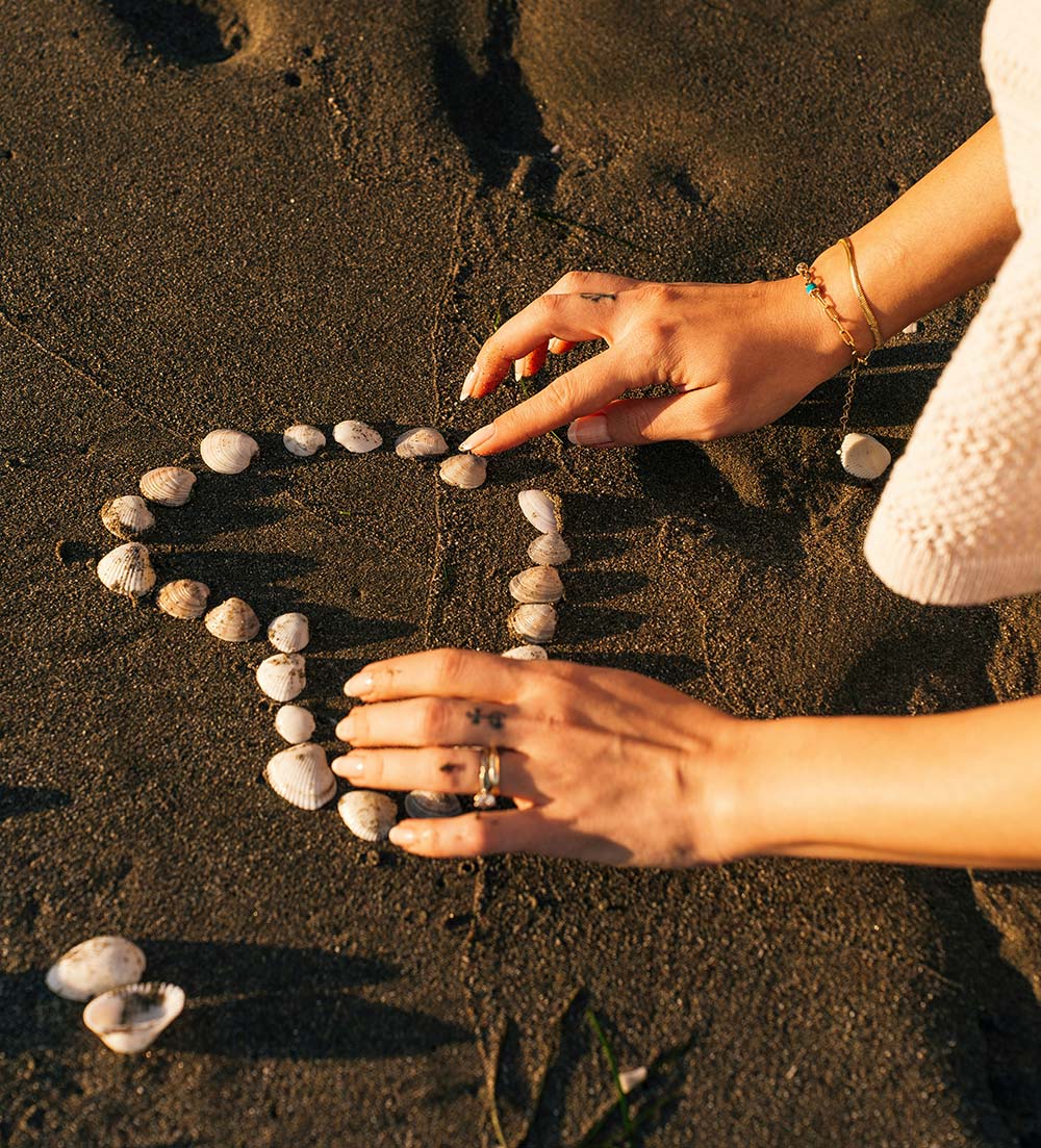 person arranging seashells in a heart shape on the sand.
