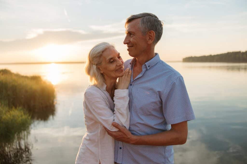 Older couple embracing by a lake at sunset symbolizing emotional closeness and intimacy in a long-term relationship