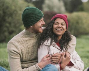 smiling couple in beanies hugging while sitting on the grass.