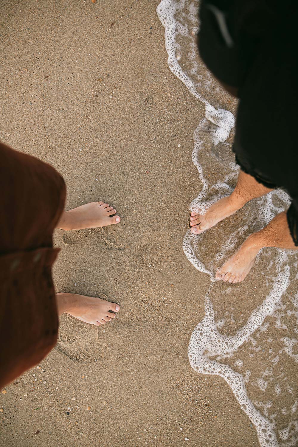top-down view of two people standing barefoot on a sandy beach as waves come in.